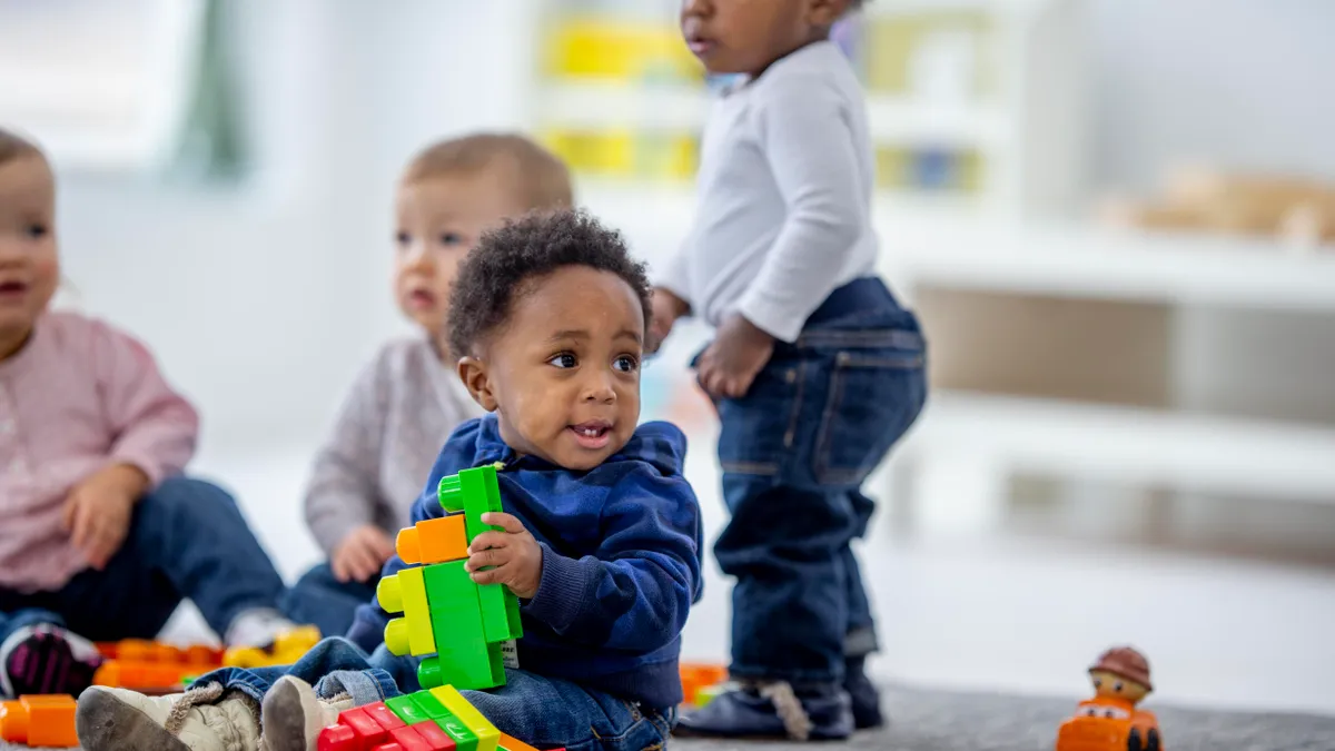 Three toddlers are pictured sitting on the ground, while one is standing. The toddler in the center is looking off to the side and is holding a plastic toy.