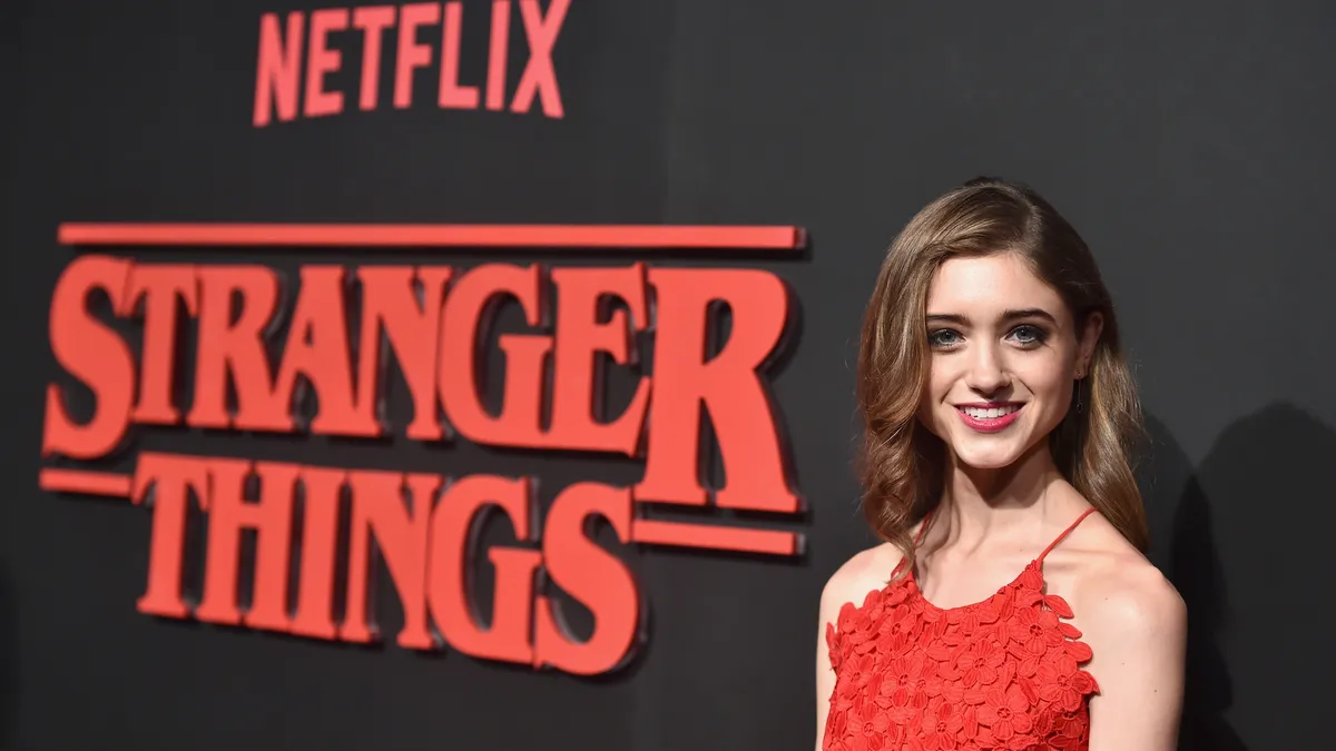 An actor in a red dress stands in front of the "Stranger Things" logo.