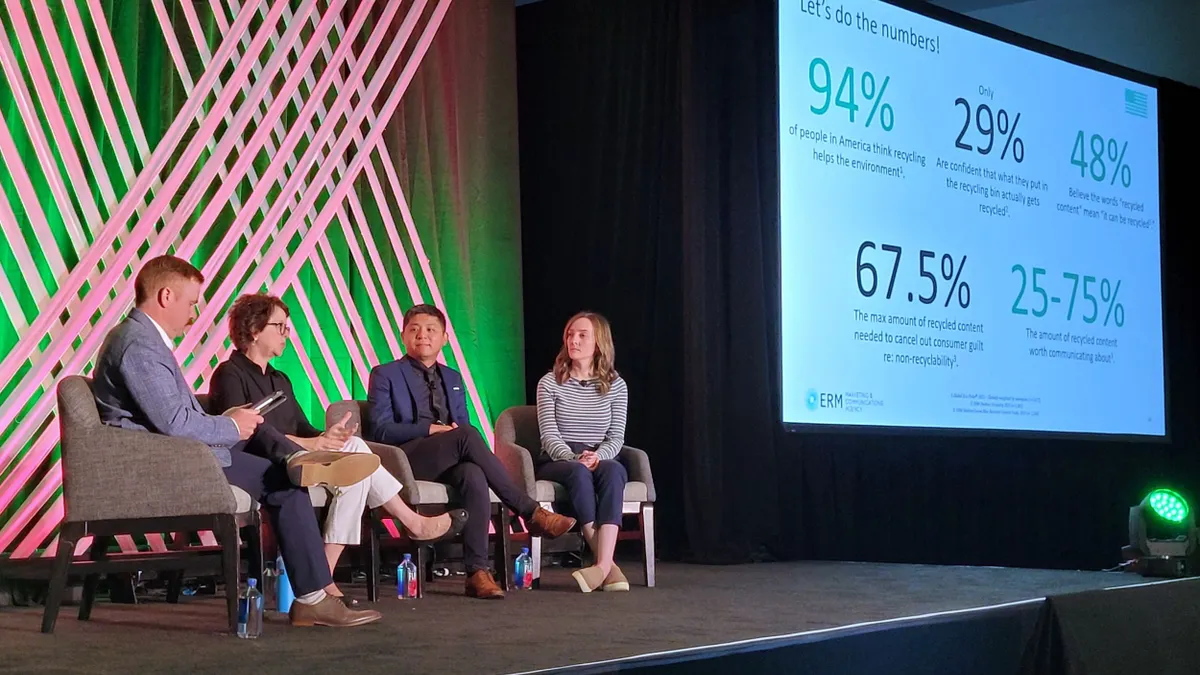 Four people sit in chairs on a stage with a large screen next to them displaying data while they address an audience.