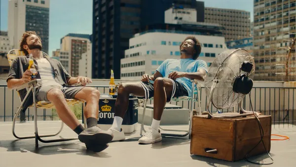 Two men relax in lawn chairs on the roof of a building, one holds a bottle of Corona.