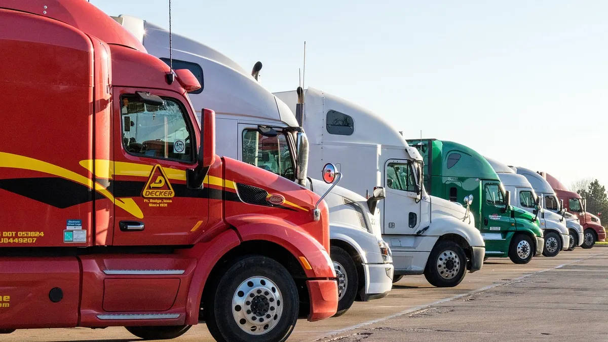 A lineup of semi trucks parked in a lot, with some trucks displaying company logos and various colors.
