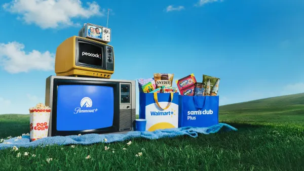 a carton of popcorn, televisions and full shopping bags on a picnic blanket on a field of grass, against a blue sky.