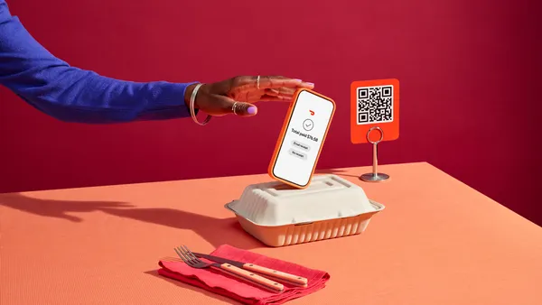 An arm holds a smartphone over a takeout box that is on a red table. On the right is a QSR code.