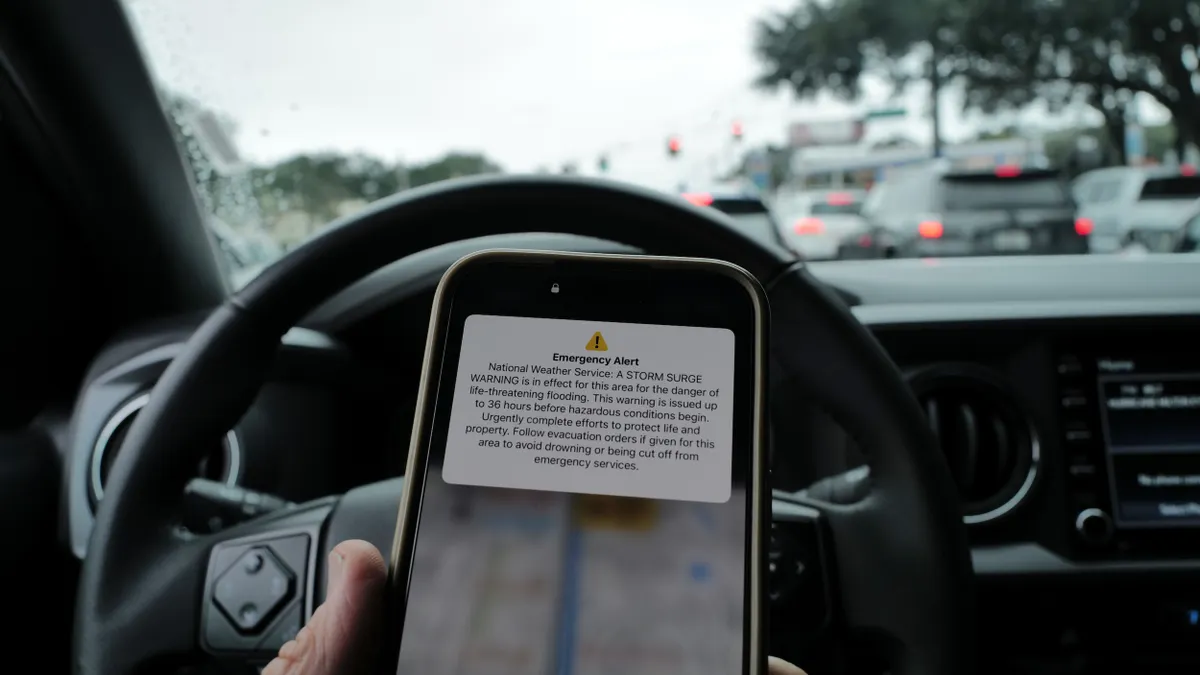 A phone showing a severe weather alert in front of a steering wheel with traffic out the front window.