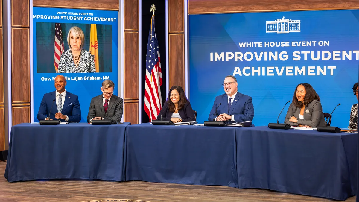 Six adults sit behind a long table. Behind them is a screen reading "White House Event on Improving Student Achievement." Another person is on a screen in the background