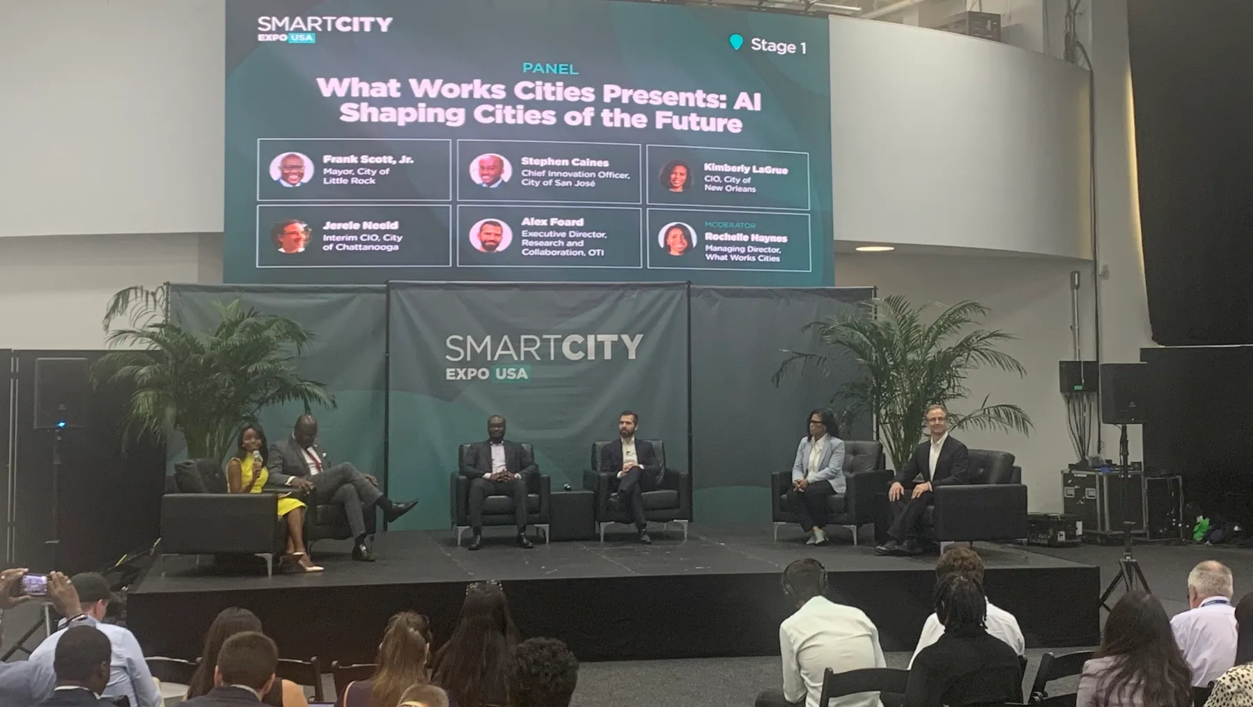 Six people sit on chairs on a stage in front of an audience. A presentation behind the speakers reads "What Works Cities Presents: AI Shaping Cities of the Future"