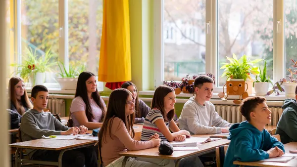 Students are sitting at individual desks in a classroom looking toward the front of the classroom