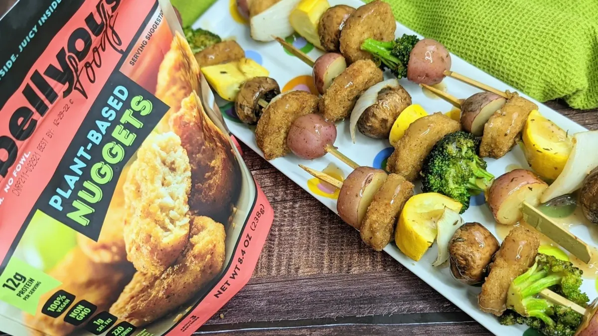 A white rectangular tray with several skewers with chicken nuggets sits on a wooden surface with a Rebellyous Foods package next to it and a green cloth behind it.