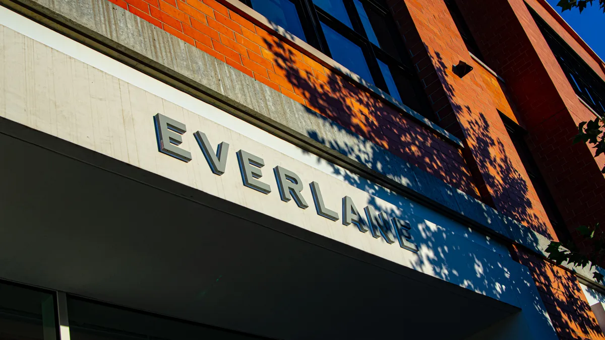 A store sign reads "Everlane" on a red brick building trimmed in concrete and covered by leafy shadows.