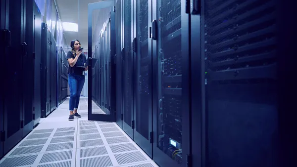 A person standing in front of a black server, holding a laptop on her left arm and speaking into her phone that's in her right hand.