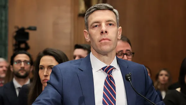Comptroller of the Currency Jonathan Gould, wearing a suit and tie, sits before a microphone during a Congressional hearing, with people sitting behind him.
