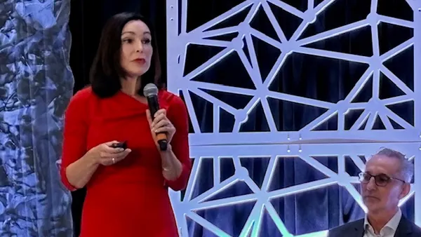 Two people are on a stage. One person is standing and has a microphone. The other is sitting.