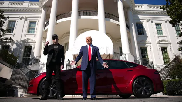 The president in a blue suit and red tie alongside Elon Musk in black with a ballcap in front of a red Tesla vehicle with the White House behind them.