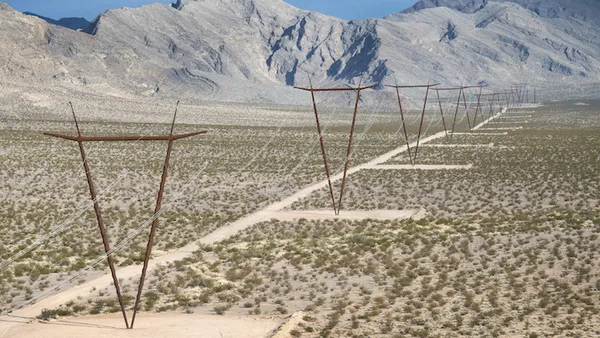 Rust-colored, V-shaped transmission towers carry power lines across a barren desert landscape with rugged, treeless mountains in the background.