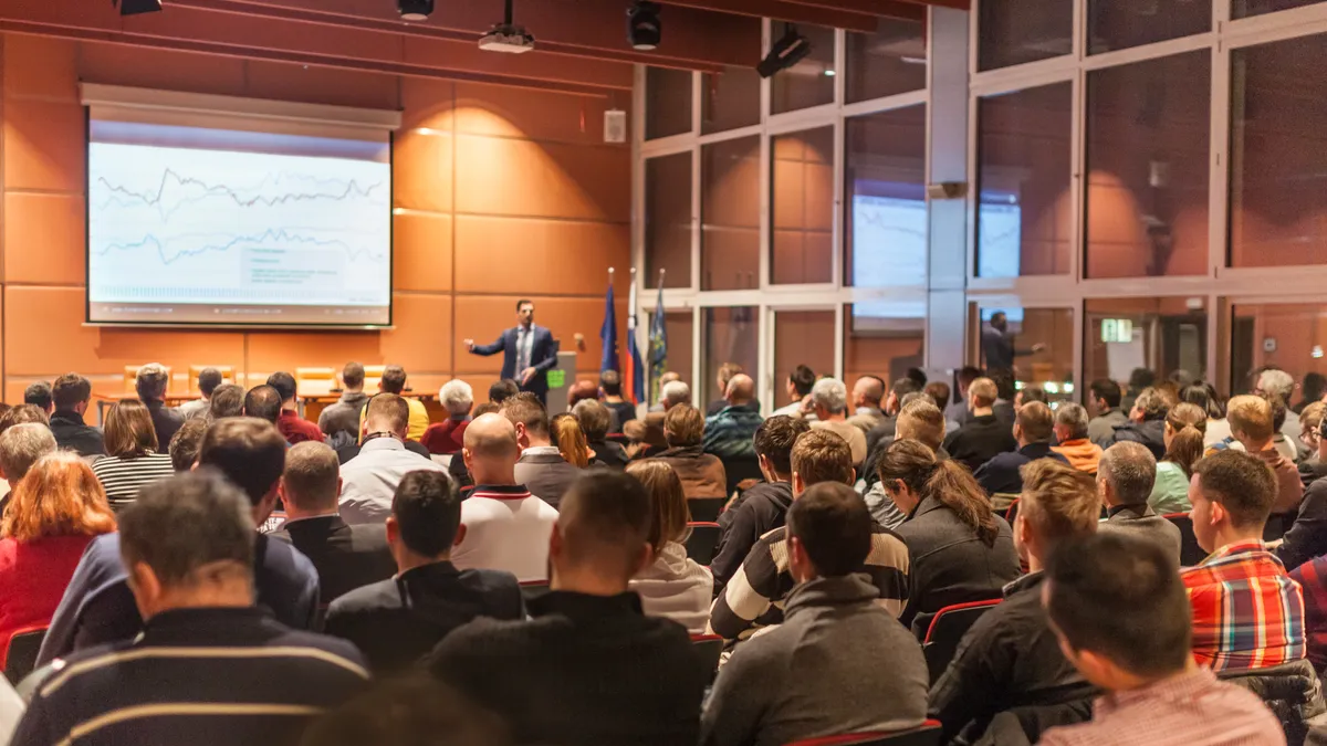 A person gives a business presentation in front of a large crown in an auditorium.