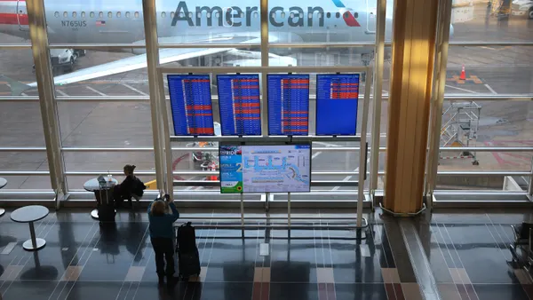 Inside an airport, a passenger stands in front of a screen displaying arrival and departure times. Behind the screen, an American Airlines aircraft on the tarmac can be seen through the window.