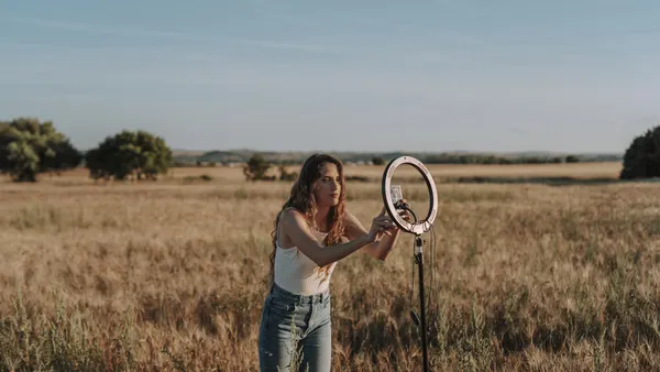 A woman in a white top and jeans sets up a ring light in the middle of a field.