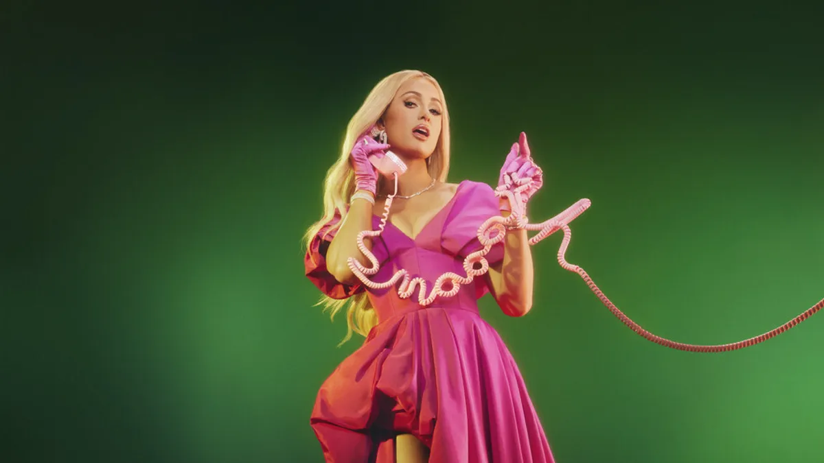 Influencer Paris Hilton wears a pink dress against an emerald green background while holding a landline phone up to her ear.