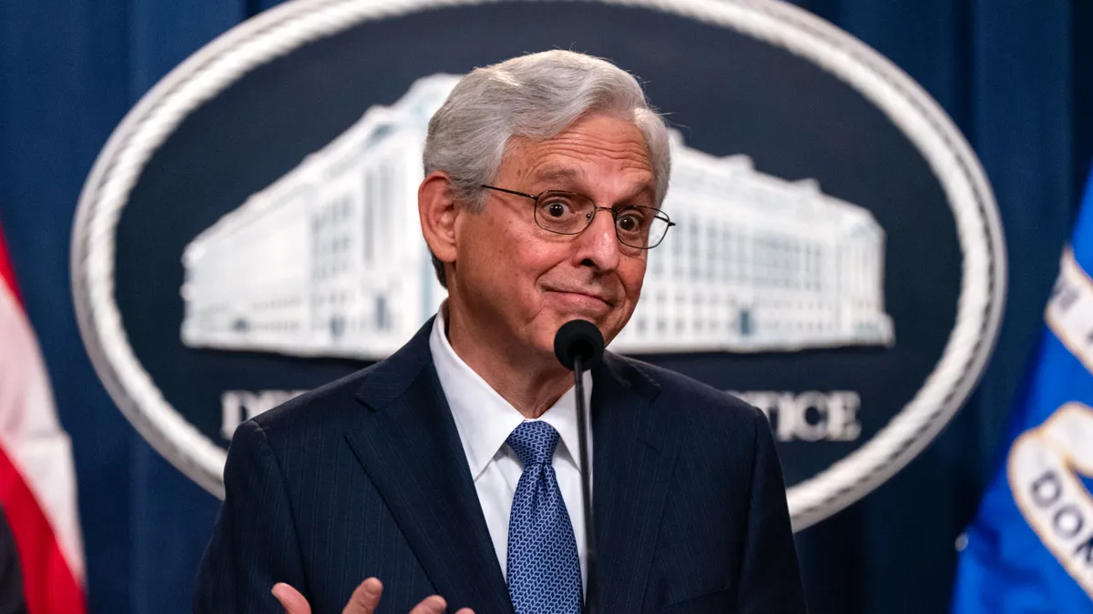 U.S. Attorney General Merrick Garland stands at a podium with hands lifted, shrugging with a smirk.