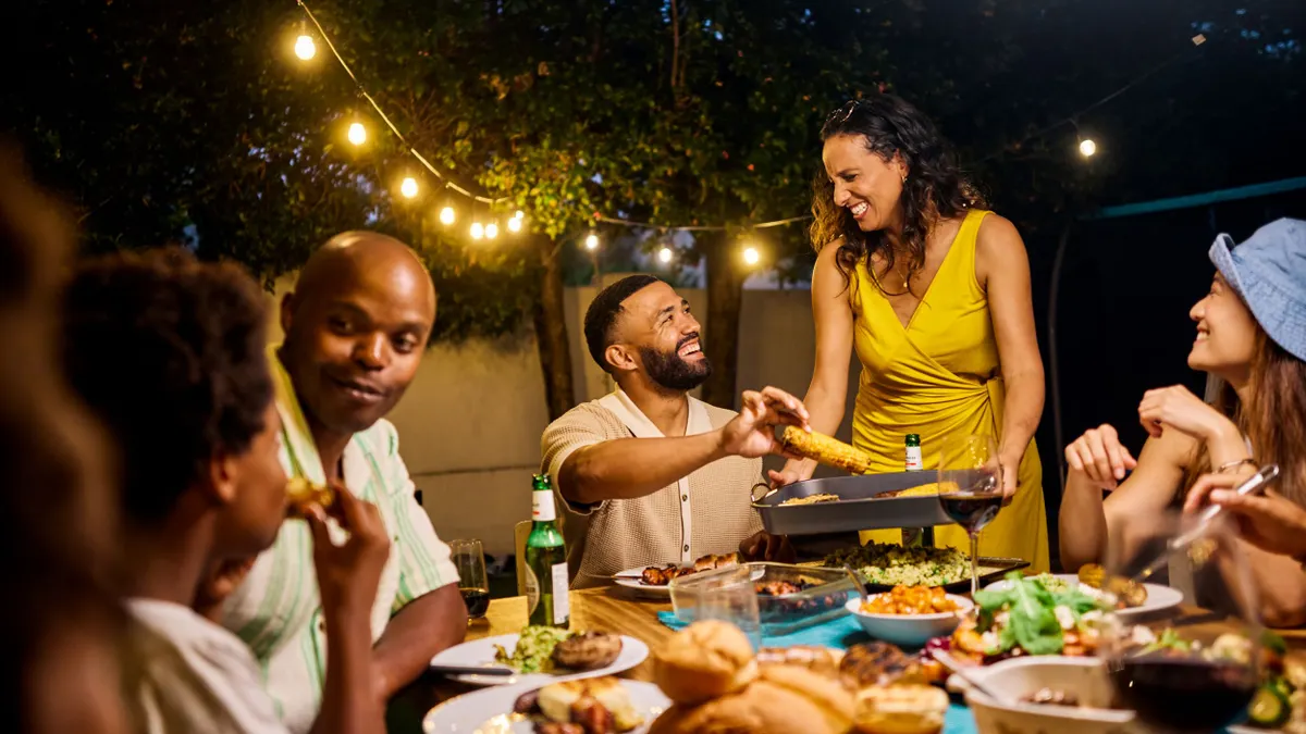 Family and friends dining at an outdoor table under string lights by the pool. Evening social gathering with food and drinks. Summer celebration concept.