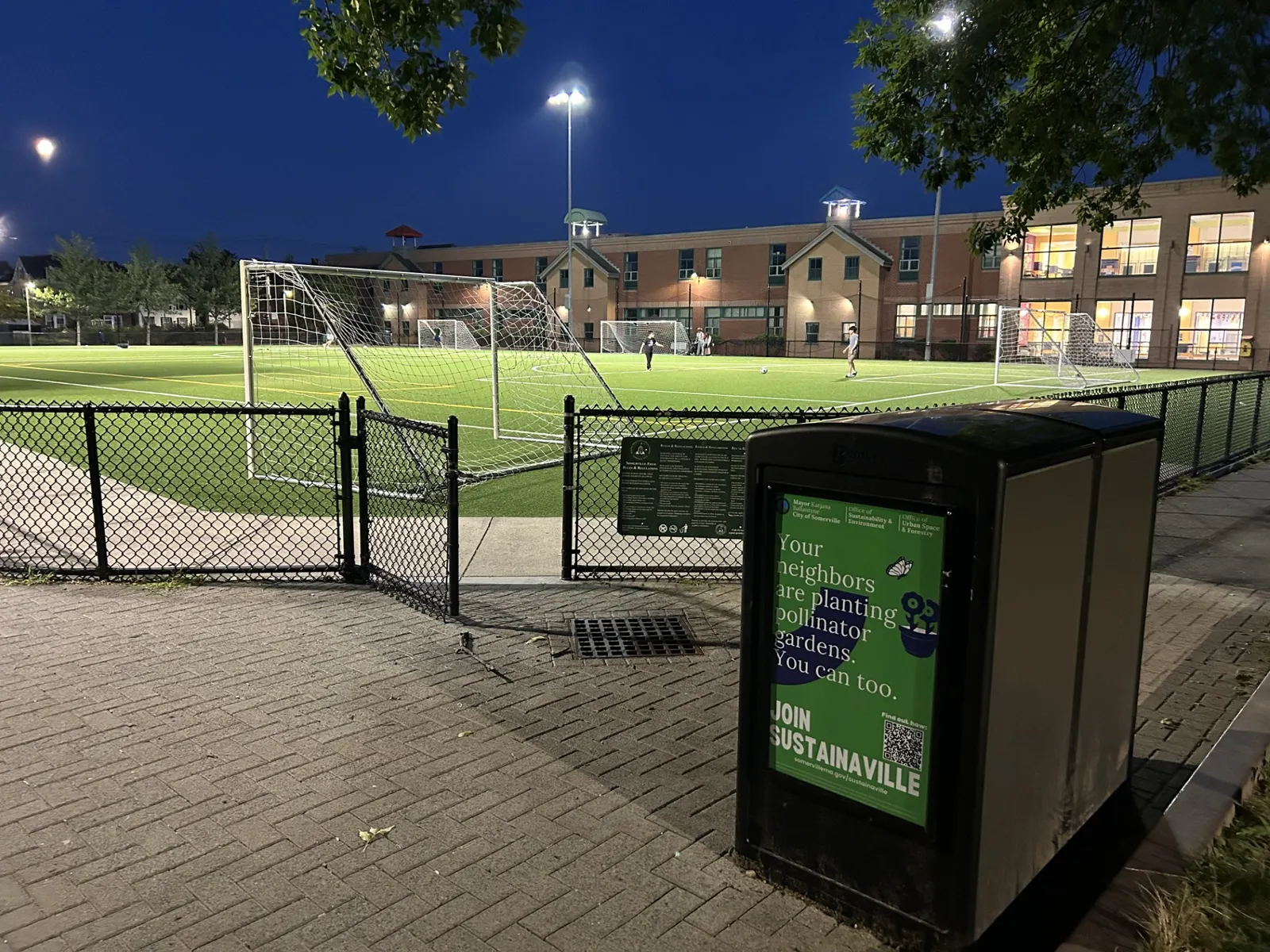 A trash can that says "Your neighbors are planting pollinator gardens. You can too. Join Sustainaville" in front of a soccer field and a school.