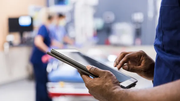 A healthcare worker in blue scrubs holds a tablet in a clinical setting