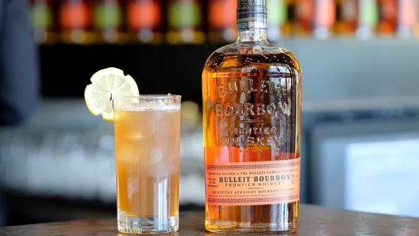 A bottle of Bulleit on a table with a blurred background. A cocktail sits next to it.