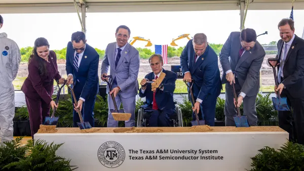Seven people in business suits and shovels scooping up dirt and smiling at the camera.