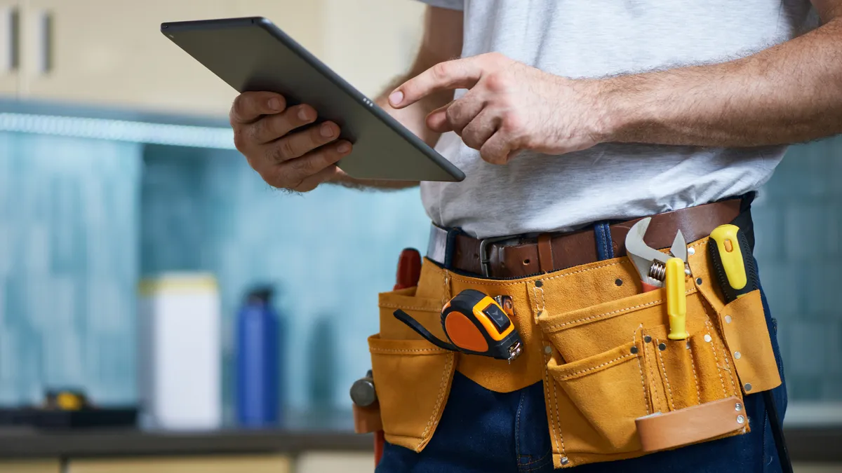 A cropped shot of a young repairman wearing a tool belt with various tools using digital tablet while standing indoors.