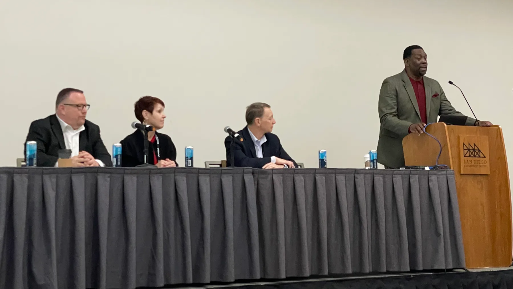 Three adult professionals are seated on a dais at a convention center table for a panel discussion as a fourth adult professional introduces them at a podium.