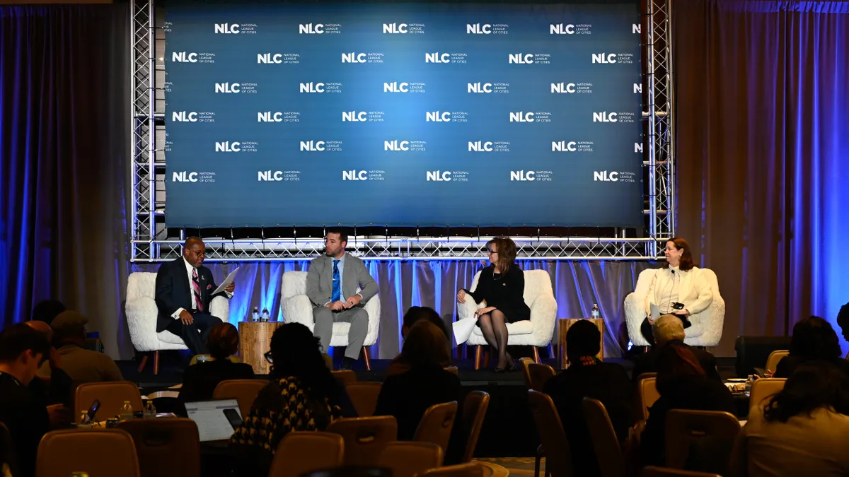 Two men and two woman seated on a stage in front of a backdrop that reads "NLC - National League of Cities."