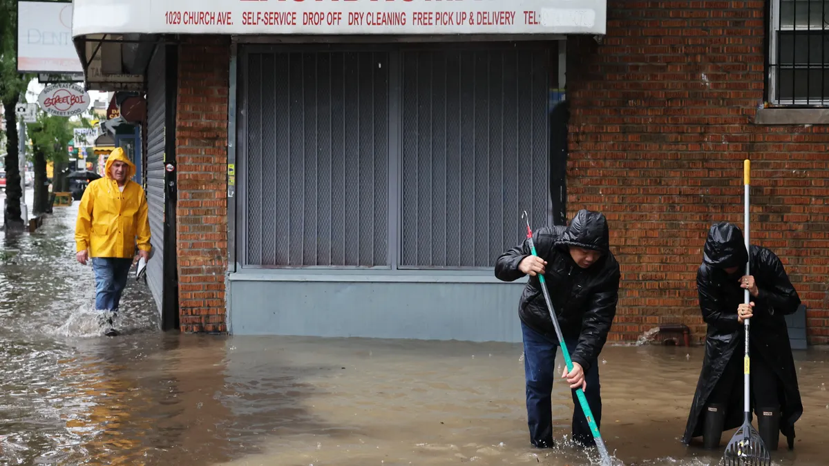 Two people holding rakes stand in floodwater up to their shins in front of a laundromat. A person in a yellow raincoat walks by.