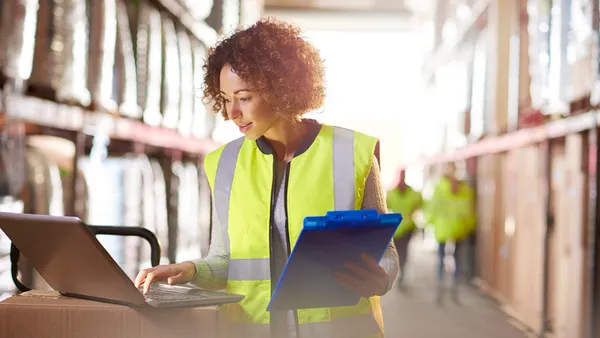 A woman wearing a yellow safety vest works on a laptop in a well-lit warehouse, holding a blue clipboard. She appears focused and diligent.