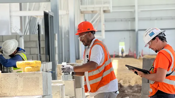 Construction workers in safety gear collaborating on a building site, with one using a tablet and others laying bricks.