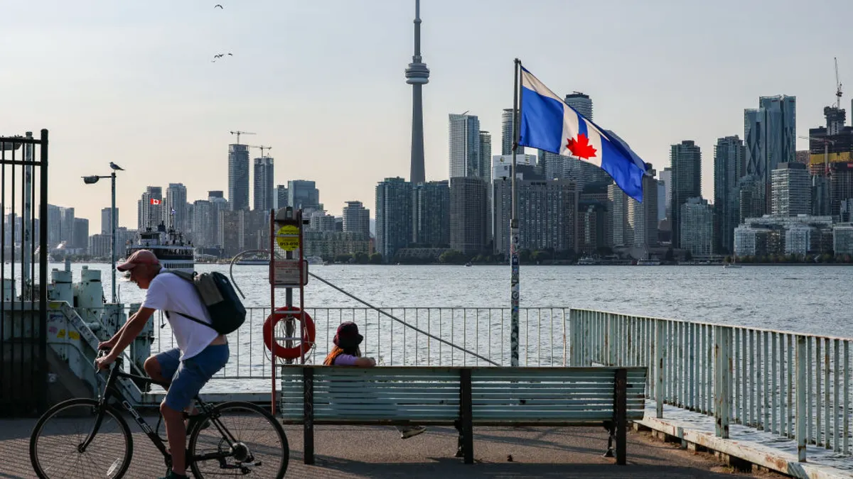 One person sits on a bicycle and another on a bench near a waterfront spot with the Toronto skyline in the distance.