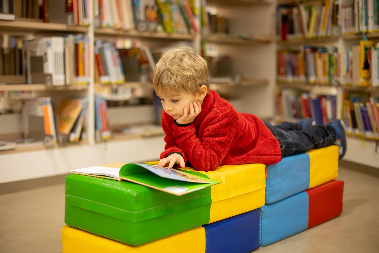 A young students lies on stomach on top of colorful cushions. The student, who is in a room with bookshelves,  is looking down at a picture book.