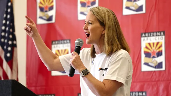 A person wearing a white t-shirt points and speaks into a microphone.