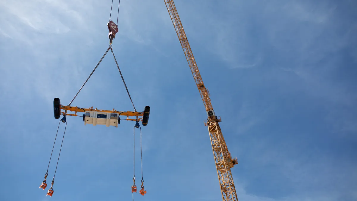 A crane carries a device in the air. It has cords that hang from the bottom.