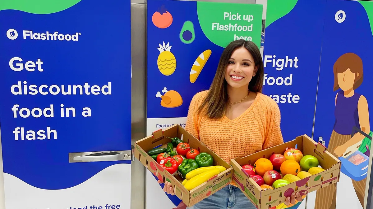 Person holding boxes of vegetables and fruits in front of blue sign that says "Flashfood get discounted food in a flash."