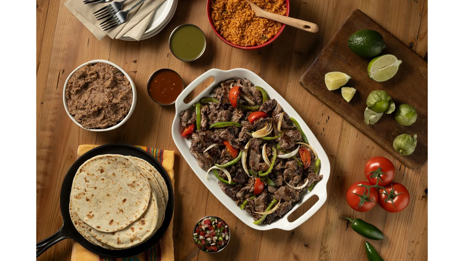 A photo of beef with peppers, onions and tomatos in a white serving dish surrounded by tomatos, hot peppers, refried beans, tortillas and other toppings.