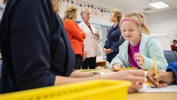 In the foreground, a teacher is working with students while they are seated at a table. In the background three people are standing and talking to each other.