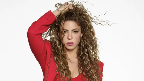 A woman in a bright red suit stands with a hand in her long, curly hair.