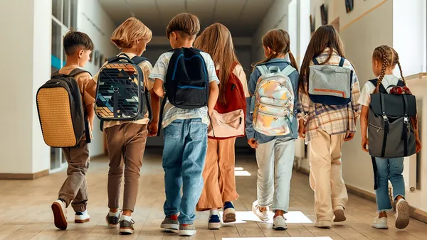 Six students carrying backpacks are walking down a hallway in a school. Their backs are to the camera and light is coming in through a window.