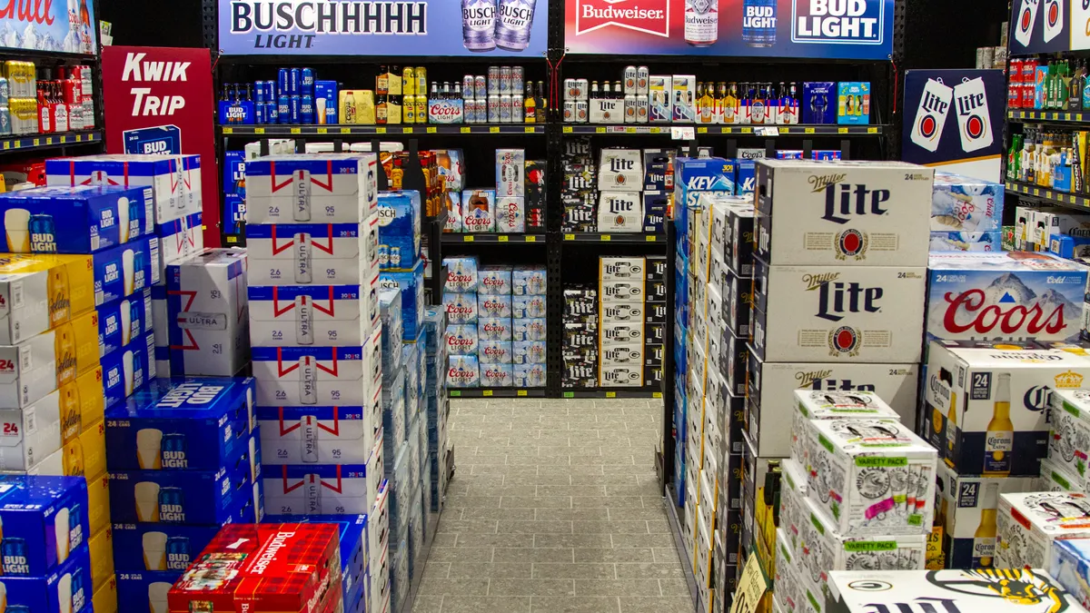 A photo of stacks and shelves of beer cases inside a room with black walls. The beer cases say things like Coors, Miller Light, Bud Light and Ultra, while signs above the shelves say Bud Light, Buschhhhhh Light and Chill.
