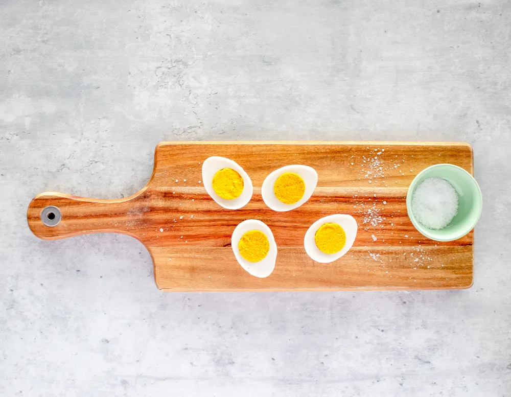A wooden cutting board with four WunderEgg halves sits on a light gray marble surface. A light green bowl with salt is to the right, and there is salt scattered on the cutting board.