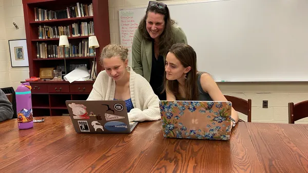 Two students sit at a table in a room looking at open laptops on the table. An adult is standing between them looking at the computers.