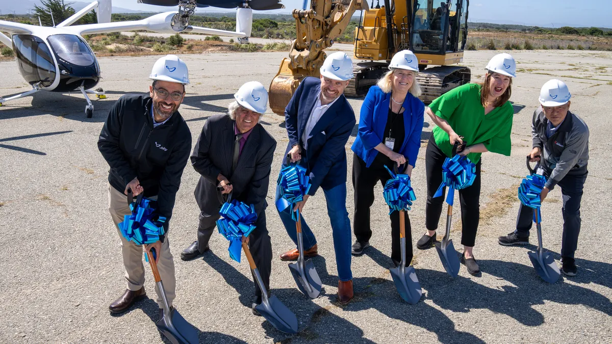 Six people wearing white hardhats and carrying shovels with a blue, shiny ribbon tied near the handles.