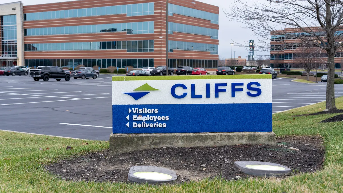 A white, blue and green Cleveland-Ciffs sign and a parking lot and a red-bricked building with windows in the background.