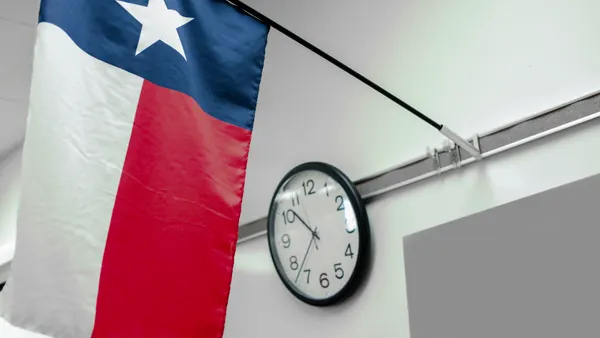 A Texas state flag hangs on a post in front of a face clock hanging in a classroom. The closeup photo shot is of the flag and clock.