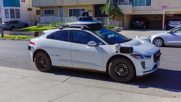 A white vehicle with sensors on the roof and sides of the car and a sign reading "Waymo" next to a sidewalk with apartment buildings across the street.